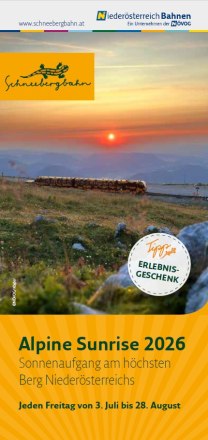 Zug der Schneebergbahn f&auml;hrt bei Sonnenaufgang &uuml;ber eine Bergwiese mit Felsen und Blumen im Vordergrund.